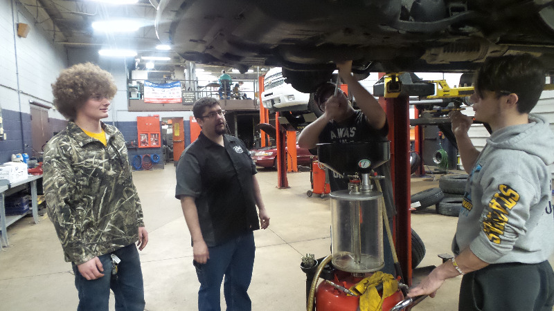 Several young people looking at a car on a raised lift.